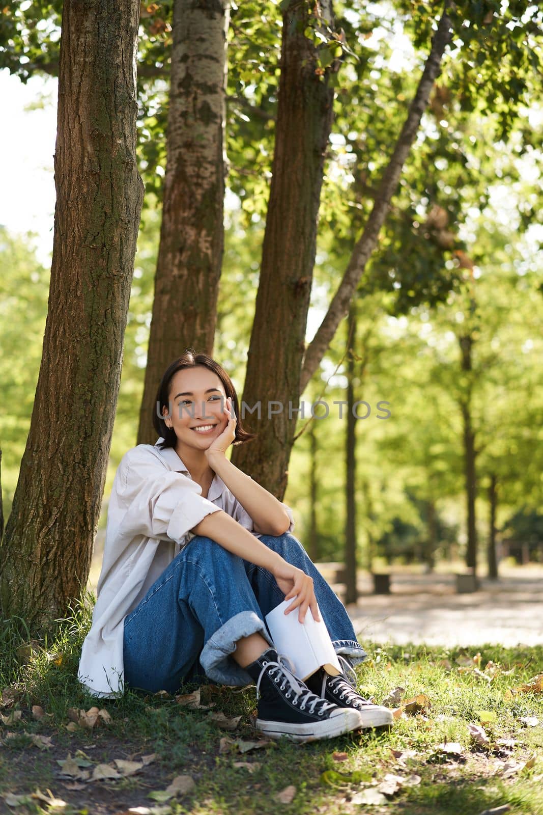 Vertical portrait of young asian woman reading her book in park, looking romantic and smiling, leaning on tree, resting outdoors.