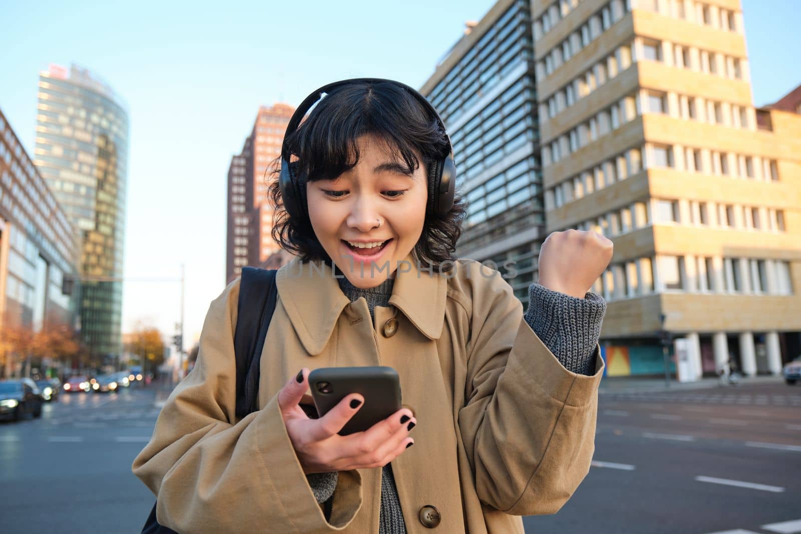 Young happy woman celebrating on street, holding smartphone and cheering, reacts amazed to good news, reads phone text message with surprised joyful face.