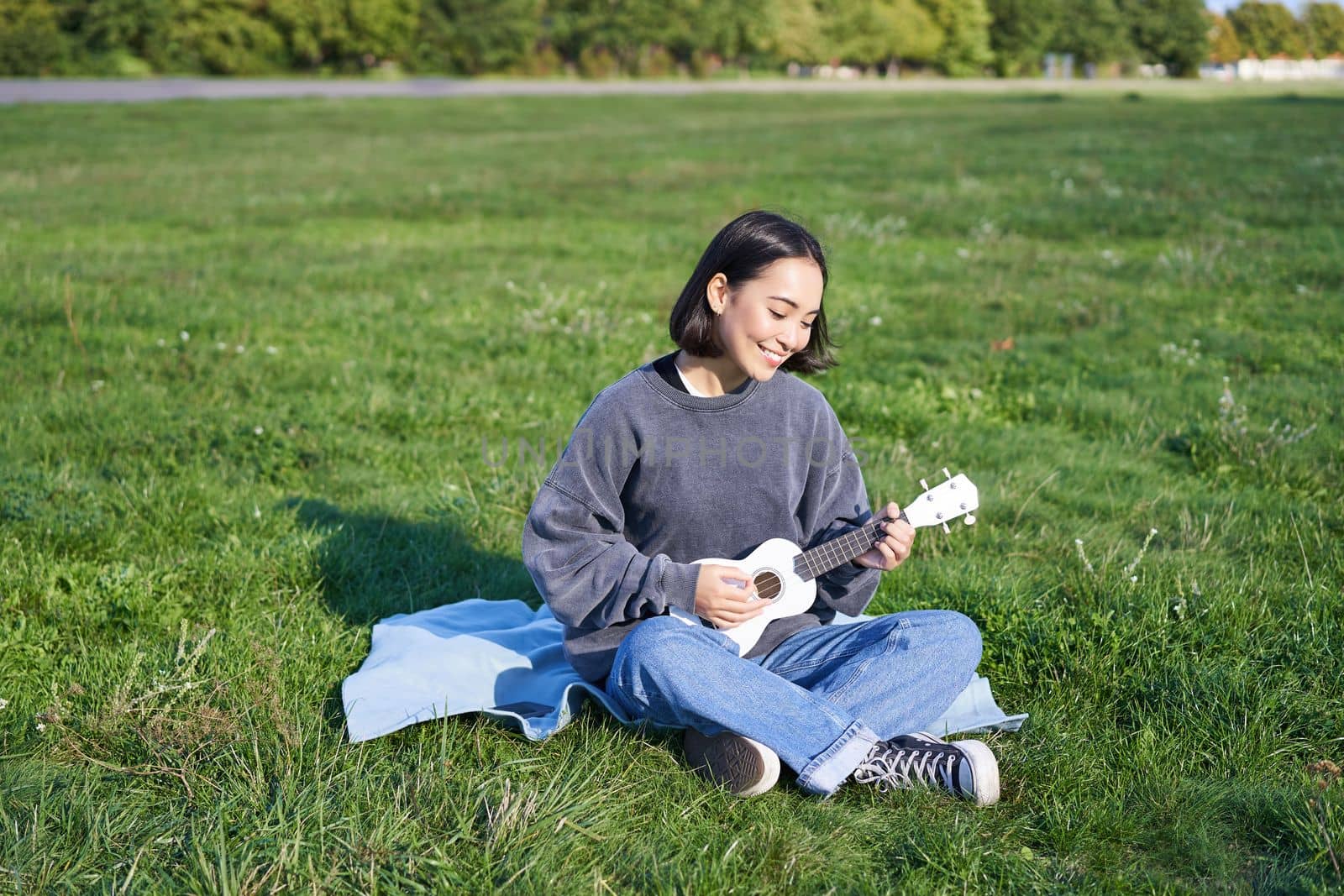 Happy brunette asian girl, student playing ukulele sitting on grass in park, relaxing, singing song, lifestyle concept.