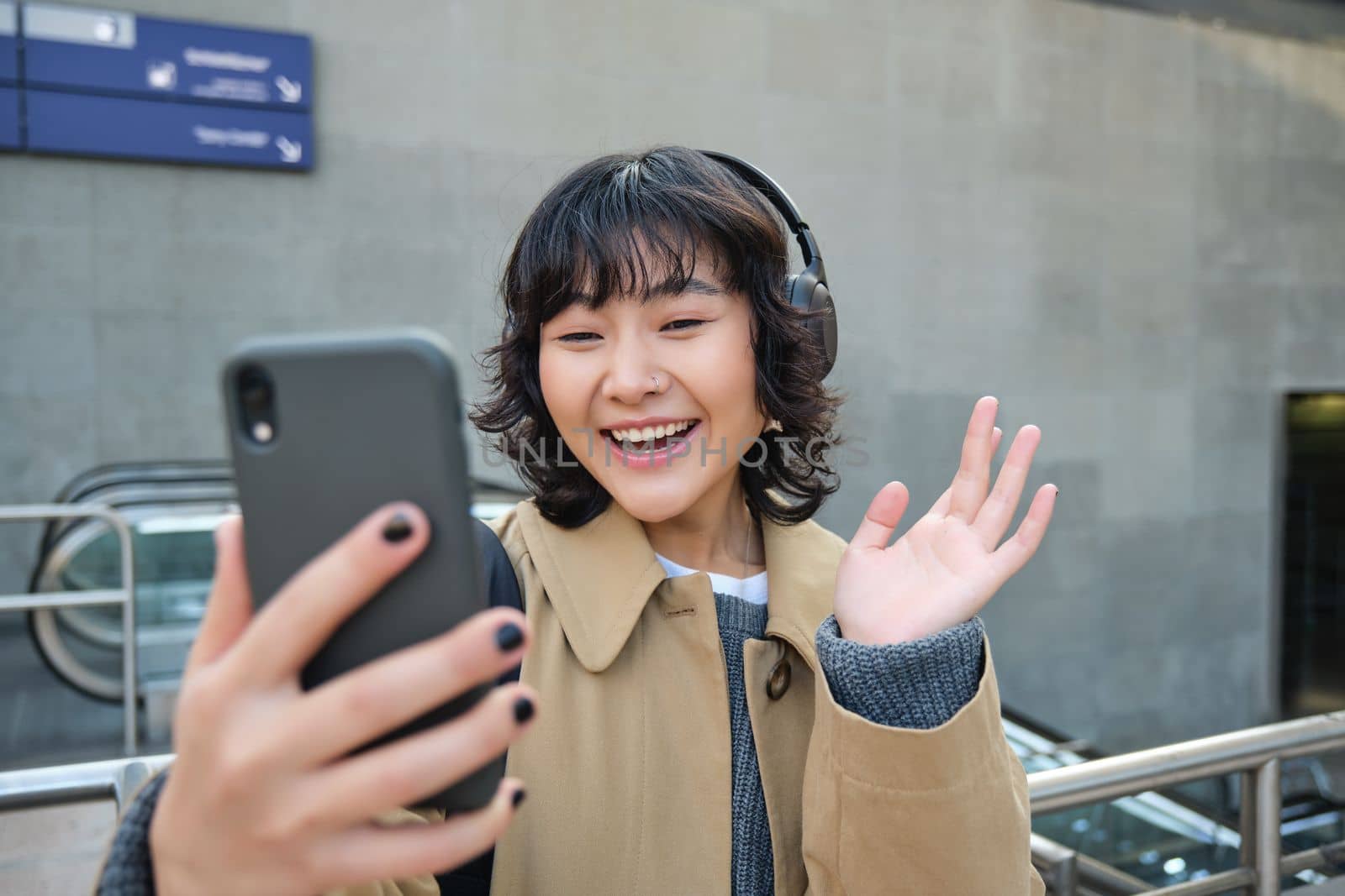 Stylish hipster girl in headphones, says hello to mobile phone camera, waves hand at smartphone, video chats, records a blog, listens music while stands on street.