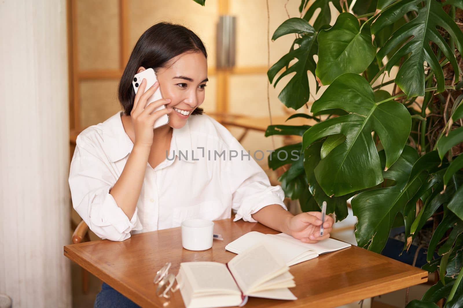 Working woman answer phone call in cafe, writing down, making notes while having conversation on telephone.