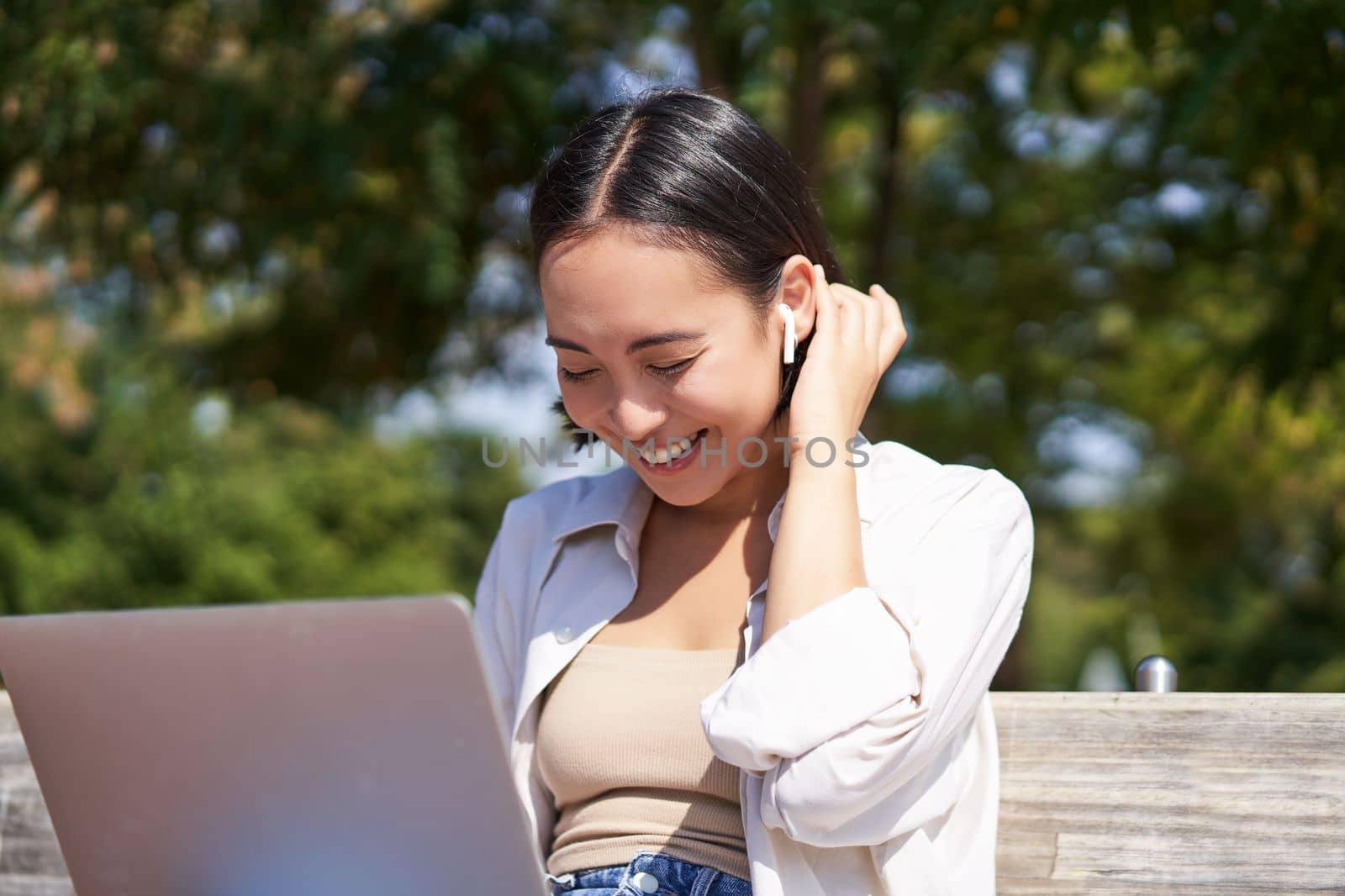 Beautiful asian girl laughing, watching video on laptop, listening music or video chatting online while sitting in park on bench by Benzoix