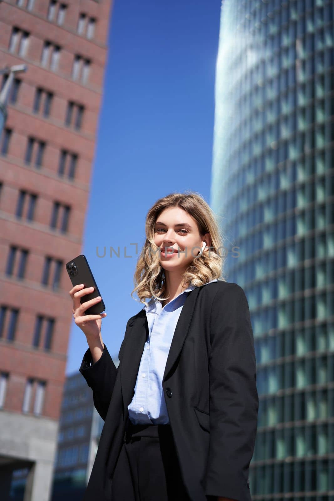 Portrait of successful businesswoman having a telephone conversation in wireless headphones, walking on street. Corporate woman with smartphone in city center by Benzoix