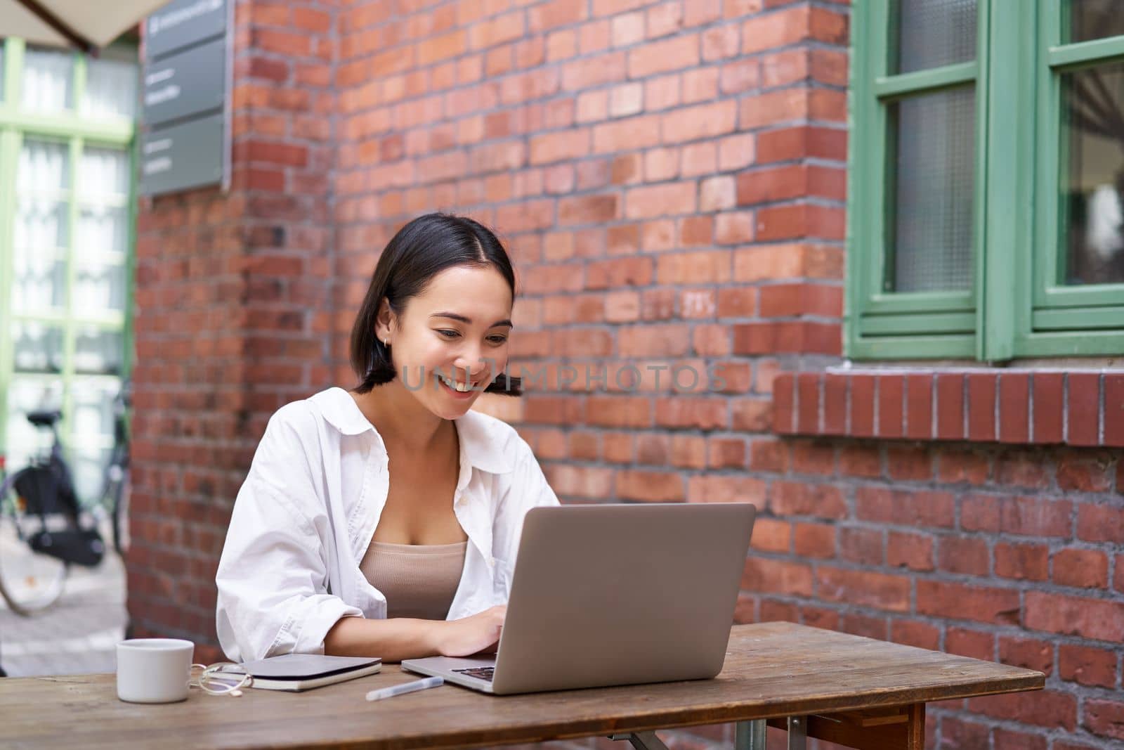 Happy asian girl watching video on laptop, doing homework outdoor in coworking space, smiling and working, drinking coffee.