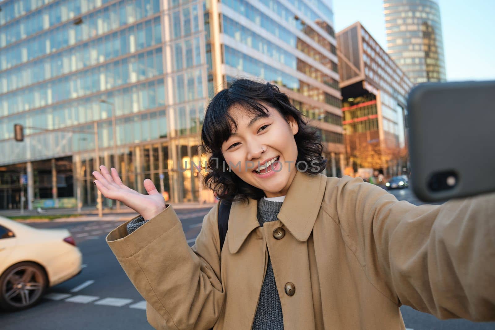 Smiling korean girl showing city, taking selfie in front of buildings on street with happy face, takes photos on her smartphone camera.