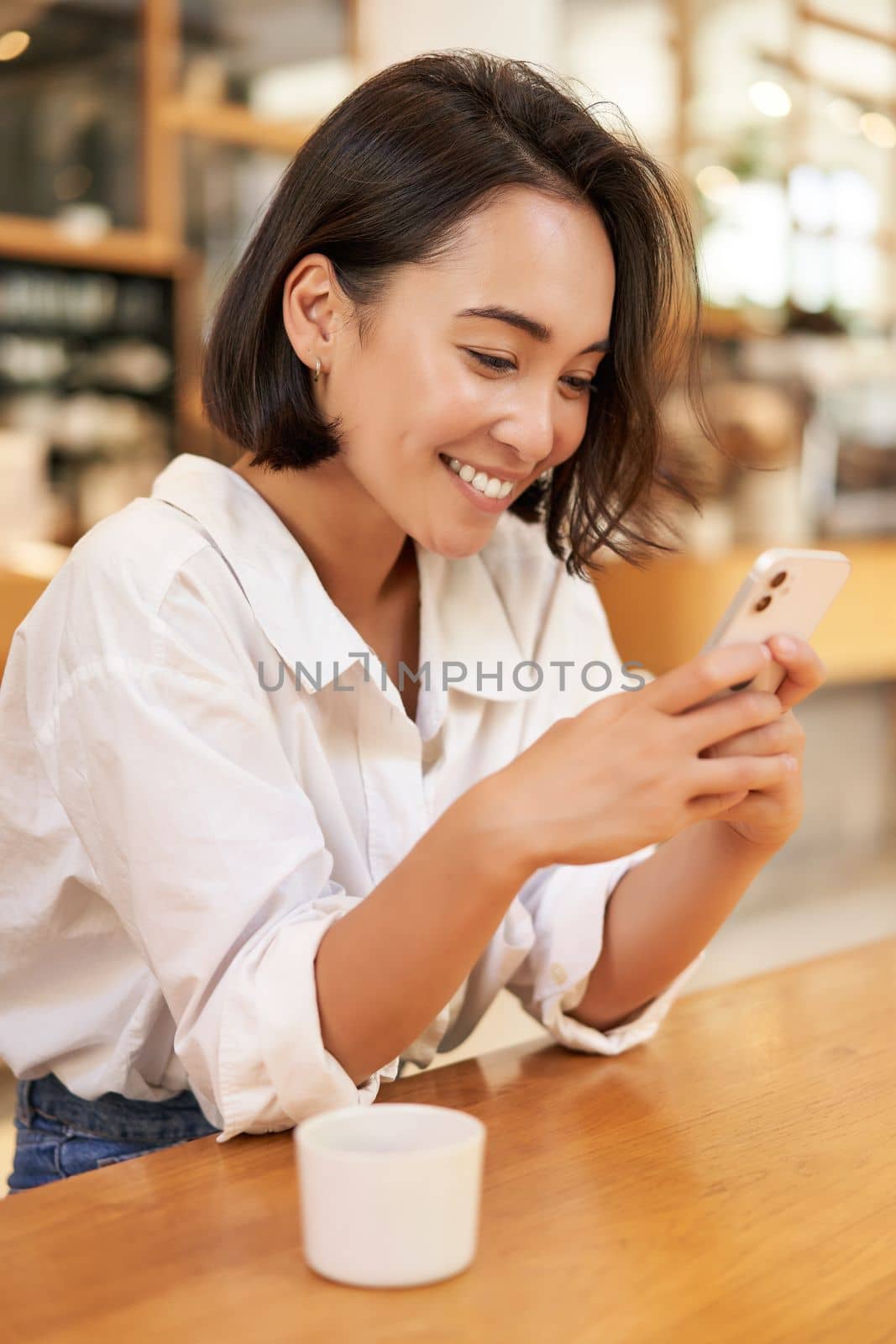 Vertical portrait of stylish asian woman sitting in cafe, drinking coffee and using smartphone by Benzoix