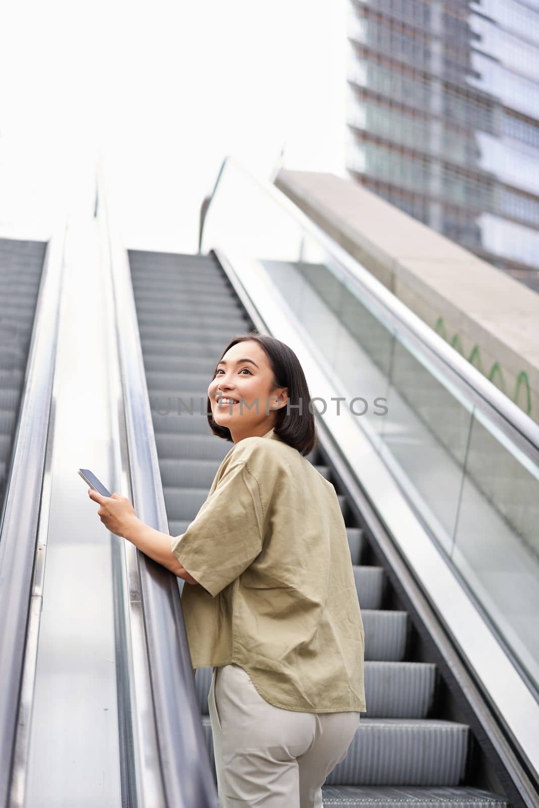 Young asian girl going up on an escalator, holding smartphone, smiling while walking in city. Copy space