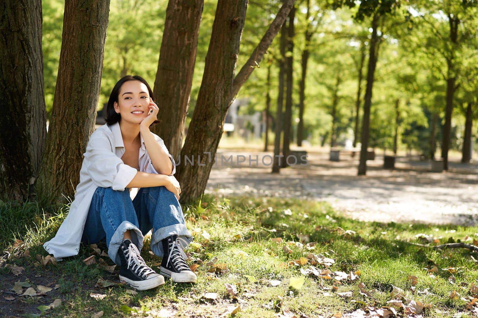 Young people. Beautiful asian girl sits near tree in park and rests, smiling and looking into distance, relaxing outdoors on fresh air in summer.