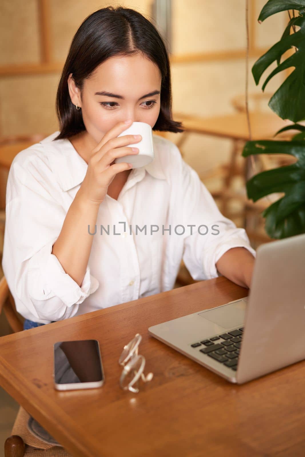 Student sitting in cafe with cup of coffee. Young asian woman working on laptop in restaurant, sitting with computer and smartphone.