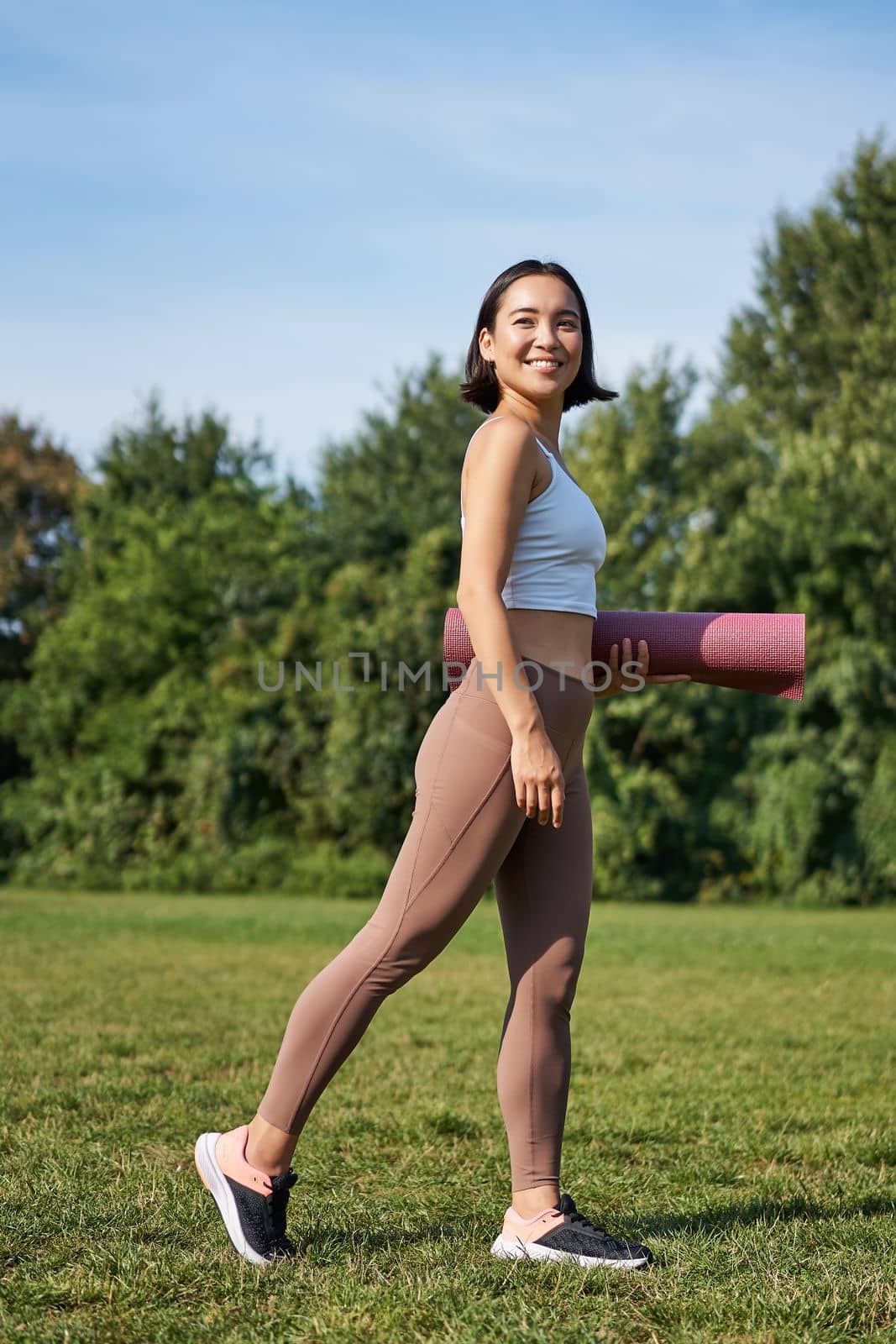 Vertical shot of stylish fitness woman with rubber mat, posing on lawn in park, doing workout on fresh air by Benzoix