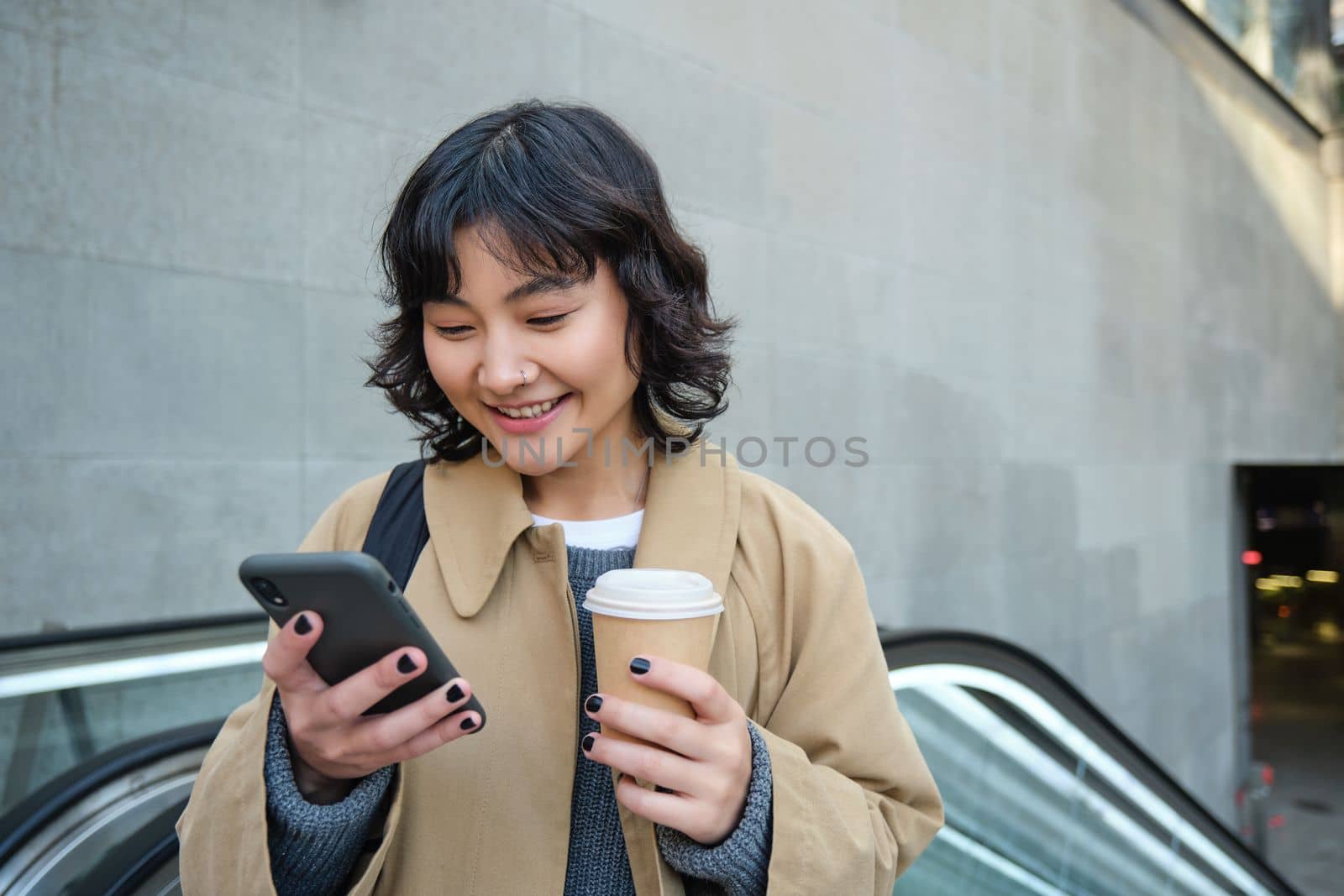 Beautiful happy asian girl, drinks coffee to go, using mobile phone while standing on escalator, walking in city centre and smiling.