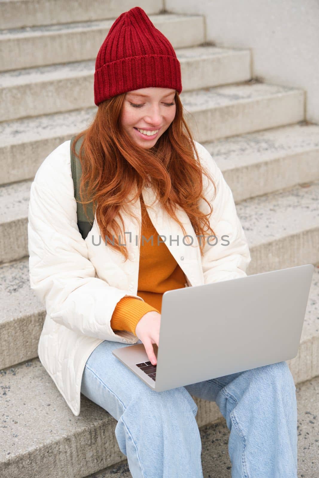 Smiling redhead girl, young woman typing on laptop keyboard, sitting outdoors on stairs with computer, working remote, doing her homework on fresh air.