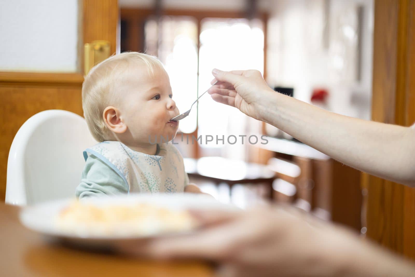 Mother spoon feeding her infant baby boy child sitting in high chair at the dining table in kitchen at home.