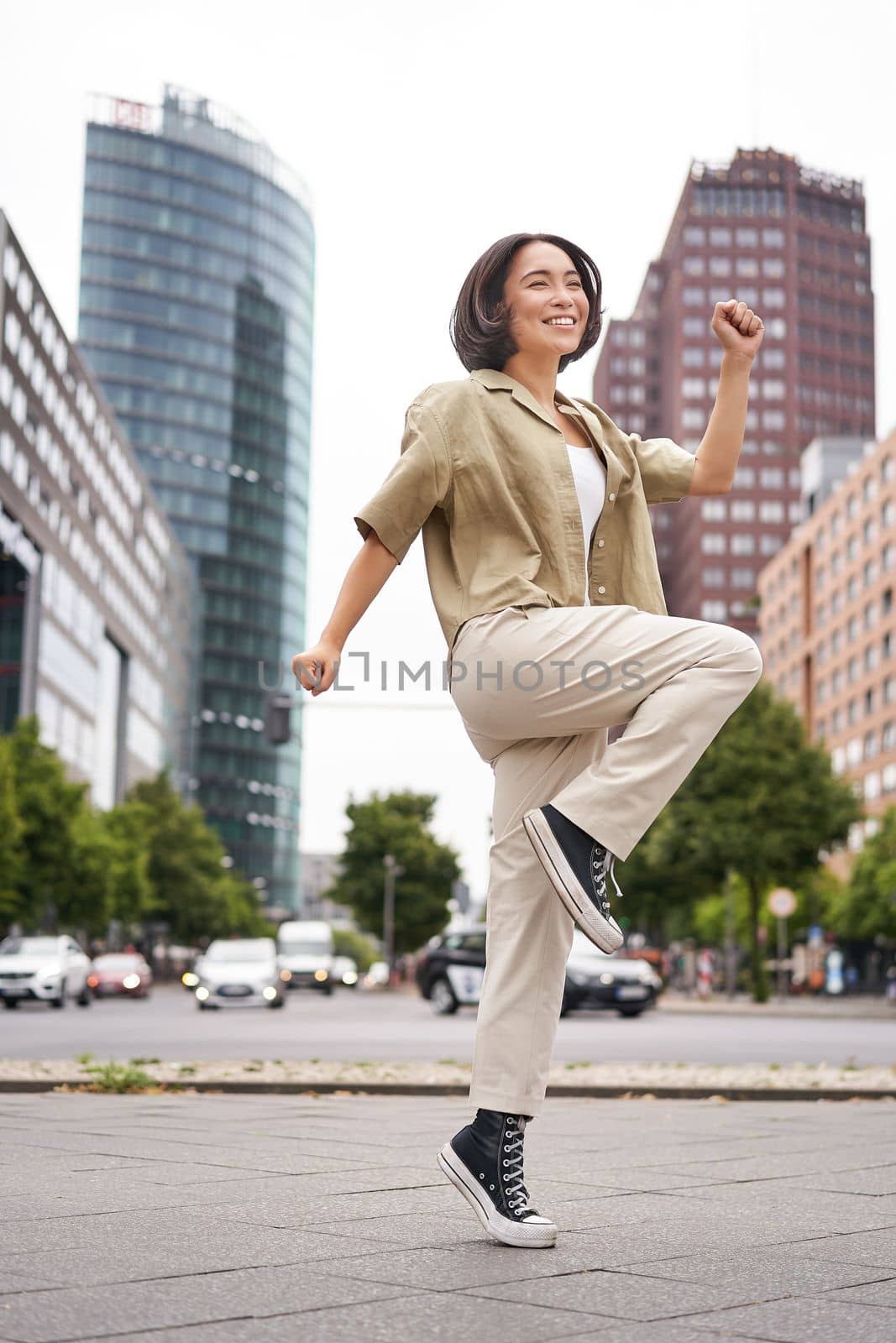 Vertical shot of young asian woman posing happy, raising hands up and dancing, triumphing, celebrating victory, enjoying day out in city by Benzoix