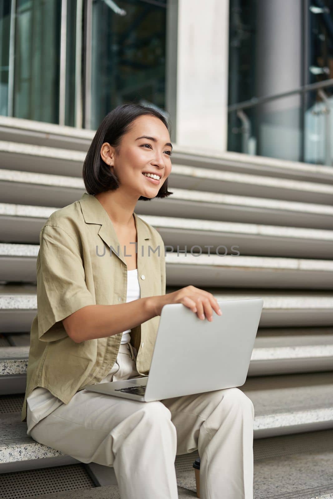 Vertical shot of smiling asian woman with laptop computer, sitting on stairs outside building. Technology and digital nomad concept