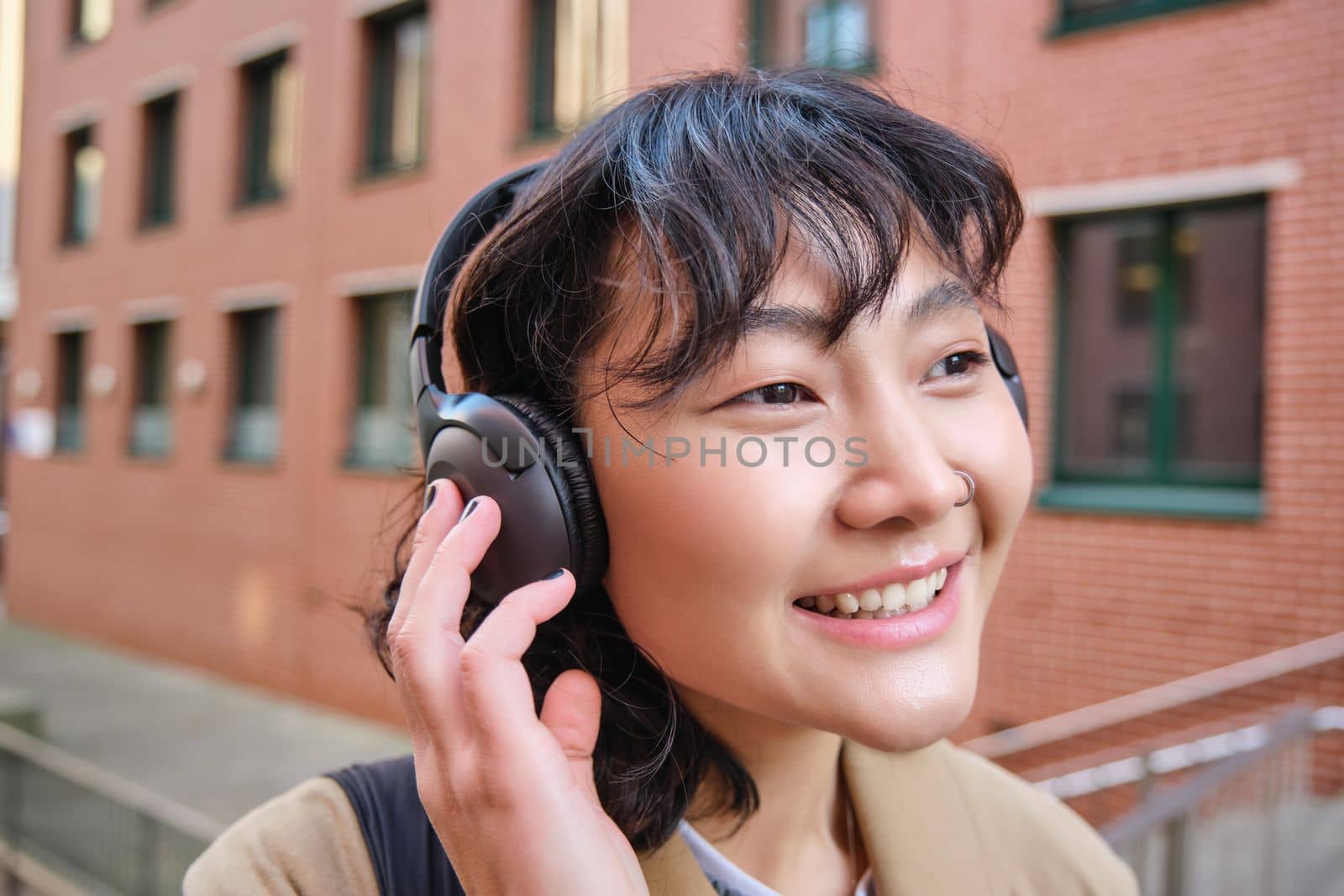 Close up portrait of stylish brunette asian girl, listens music in headphones, touches earphones and smiles, enjoys favorite song, stands on street.