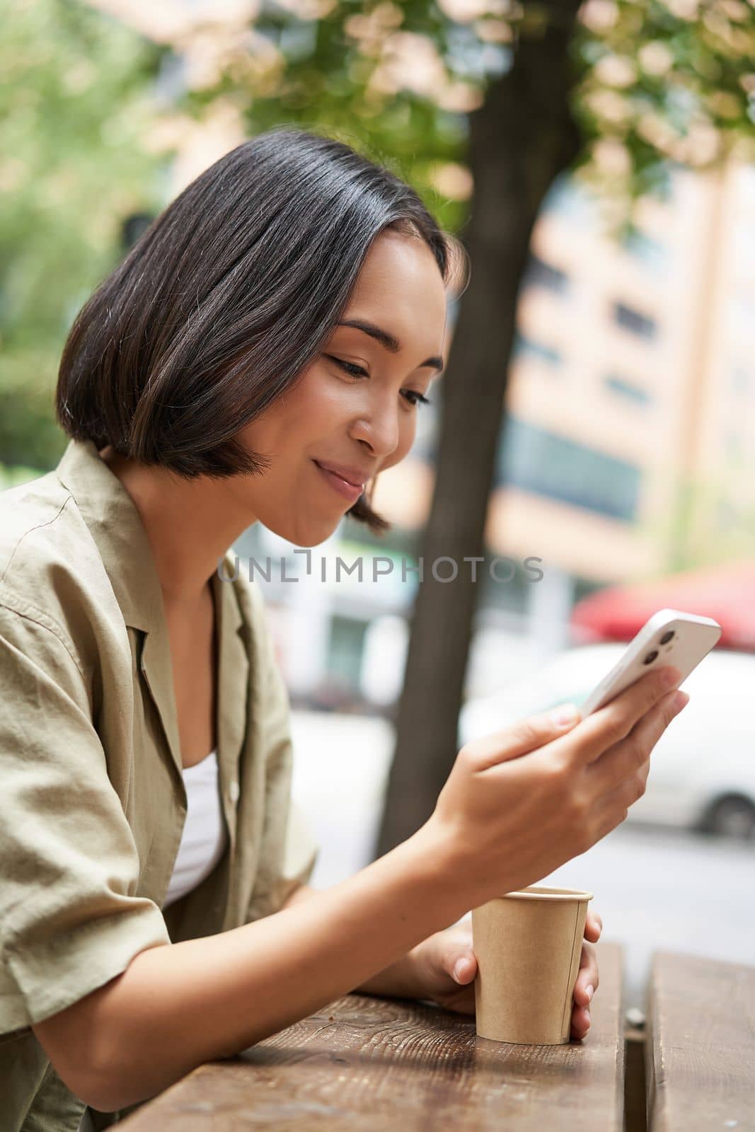 Close up portrait of asian girl sitting in cafe outside, drinking coffee and using smartphone, reading mobile phone.