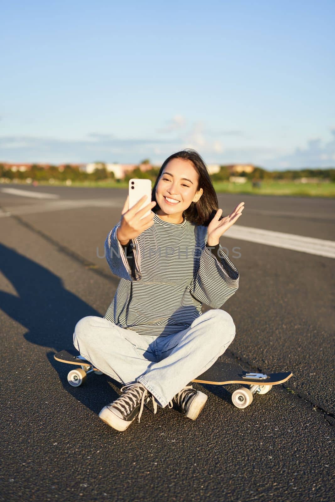 Happy teen asian girl takes selfie, video chats on smartphone app while sits on skateboard, skater records vlog while sits on longboard by Benzoix