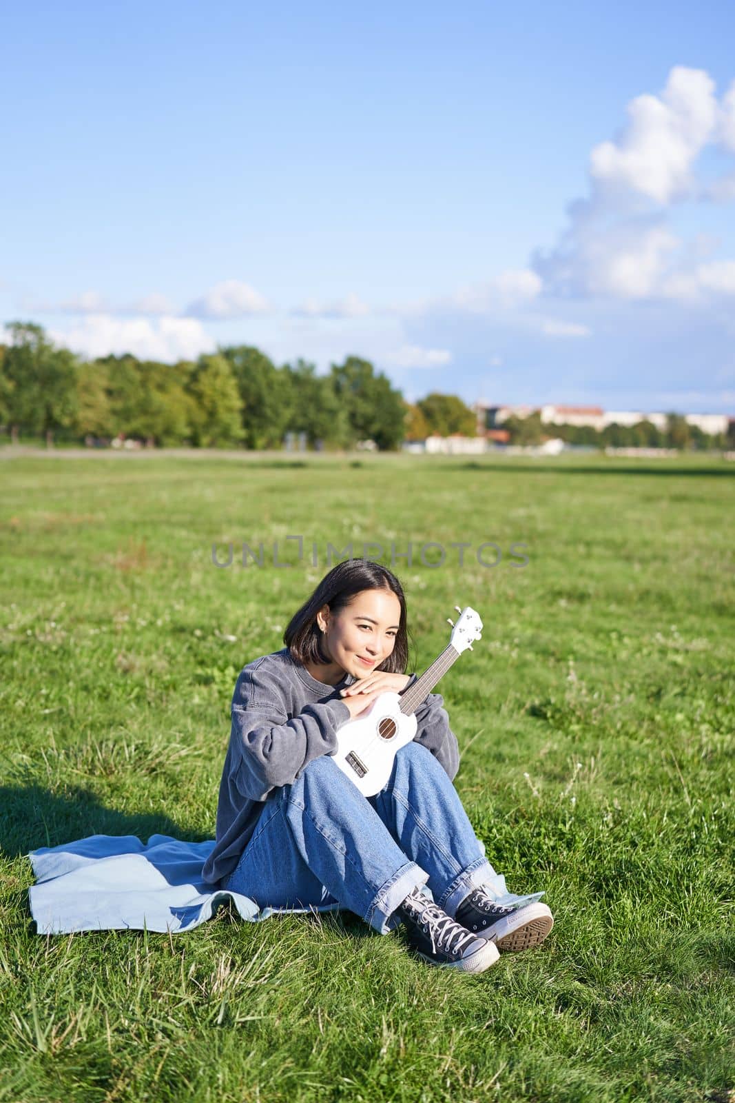 Portrait of asian girl sitting alone in park, playing ukulele and singing, enjoying leisure time. Copy space