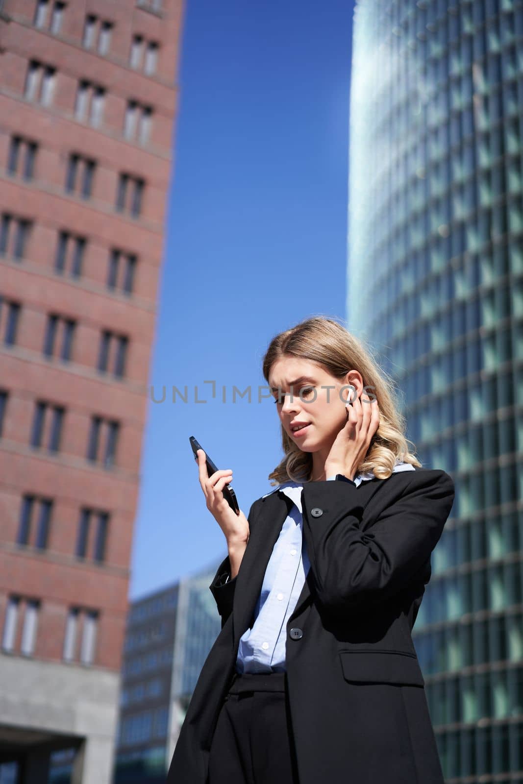 Portrait of successful businesswoman having a telephone conversation in wireless headphones, walking on street. Corporate woman with smartphone in city center.