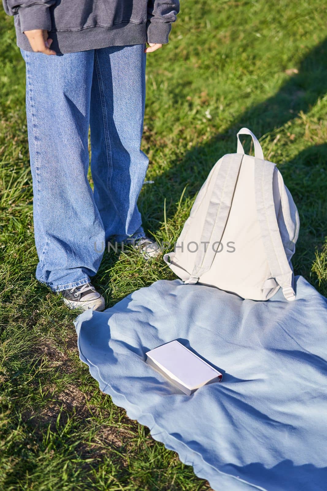 Vertical shot, young teenage girl body, standing next to blanket with backpack and book, picnic time outdoors.