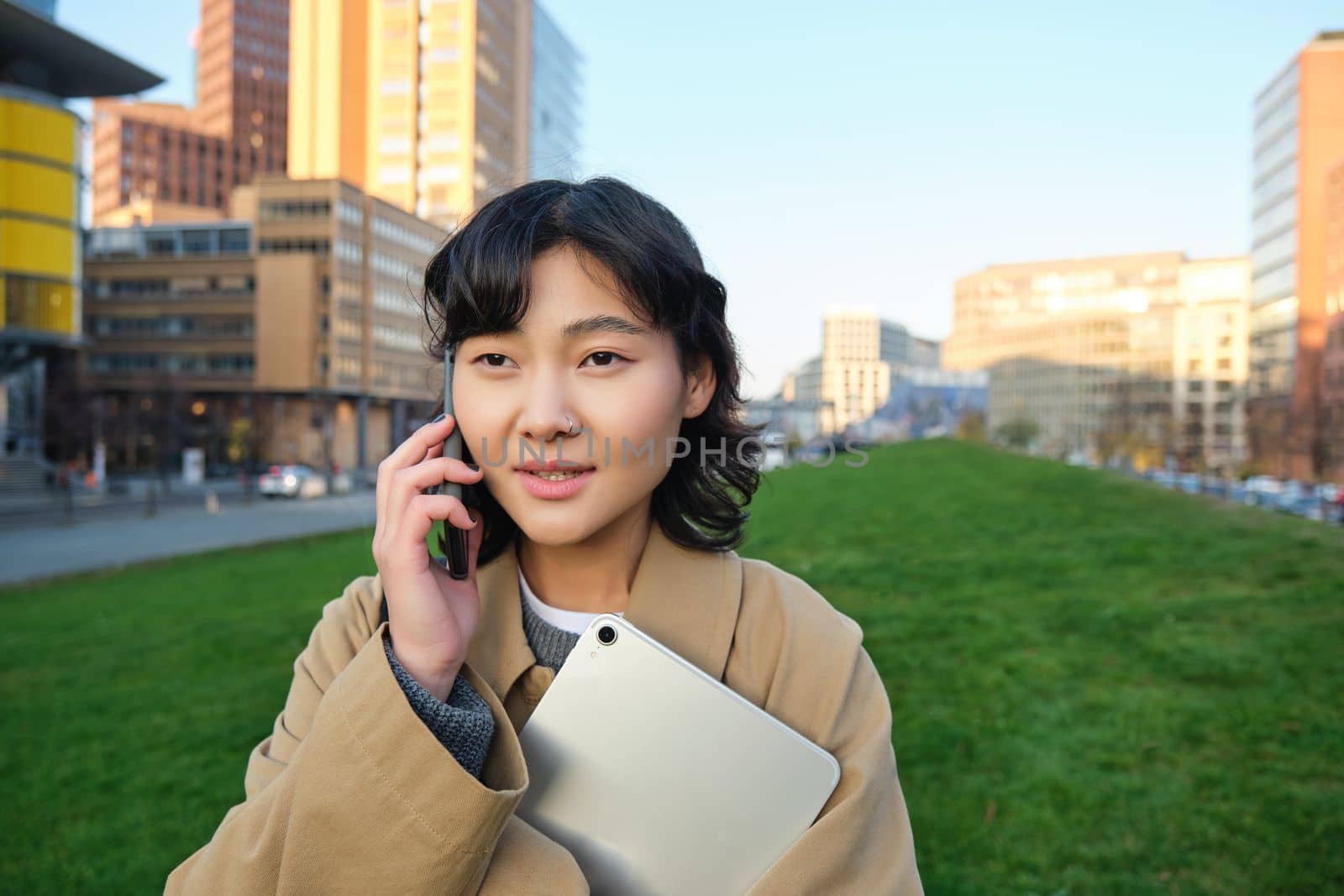 Stylish smiling girl, university student in trench coat, holds tablet, talks on mobile phone, has conversation over telephone and looks relaxed, stands on street.