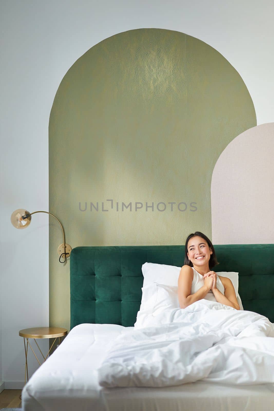 Vertical shot of beautiful asian girl lying in bedroom with sophisticated interrior, smiling and looking happy in morning, spending time in bed.