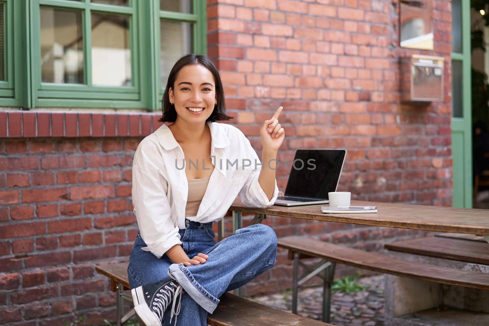 stylish modern asian girl with laptop, sitting in cafe, looking amazed and pointing at upper right corner banner, showing info advertisement.