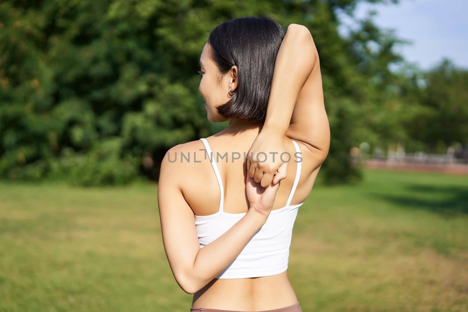 Rear view of young sporty woman stretching her arms behind back, warm-up, prepare for workout jogging, sport event in park by Benzoix