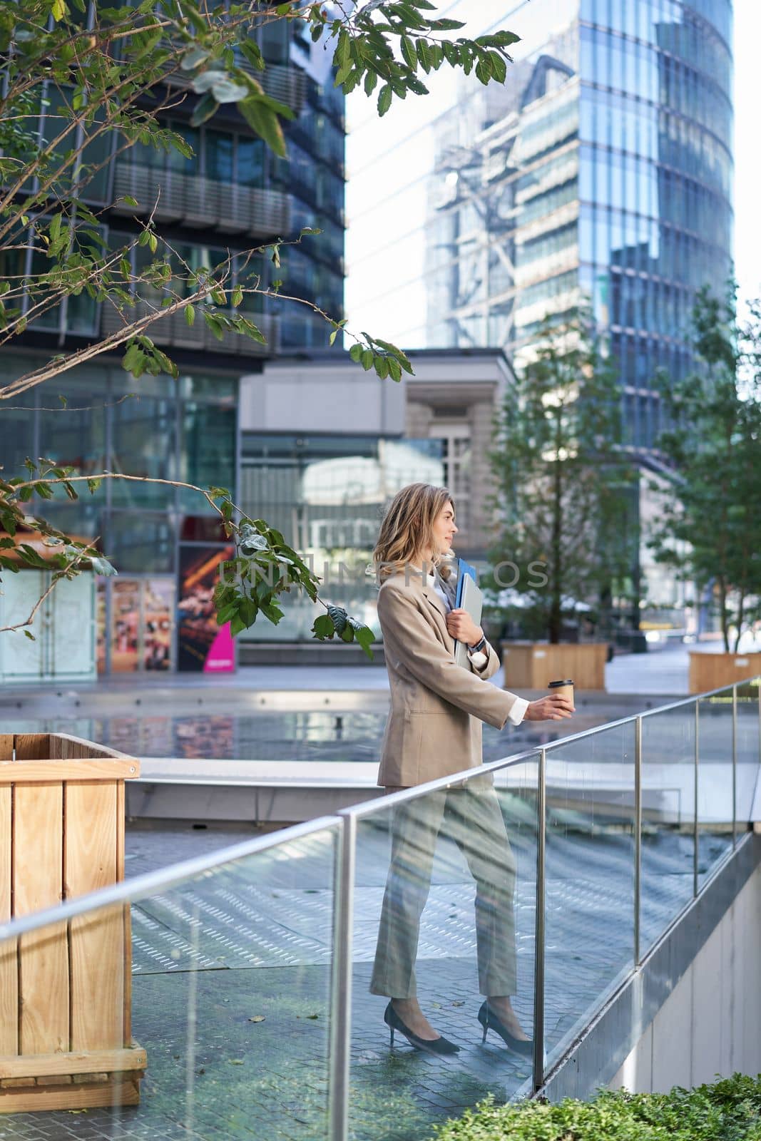 Vertical shot of corporate woman in beige suit standing outside on street, drinks morning coffee takeaway before work.