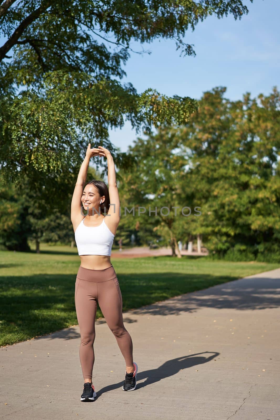 Smiling Asian girl stretching after good workout in park, listening music in wireless headphones, jogging outdoors.