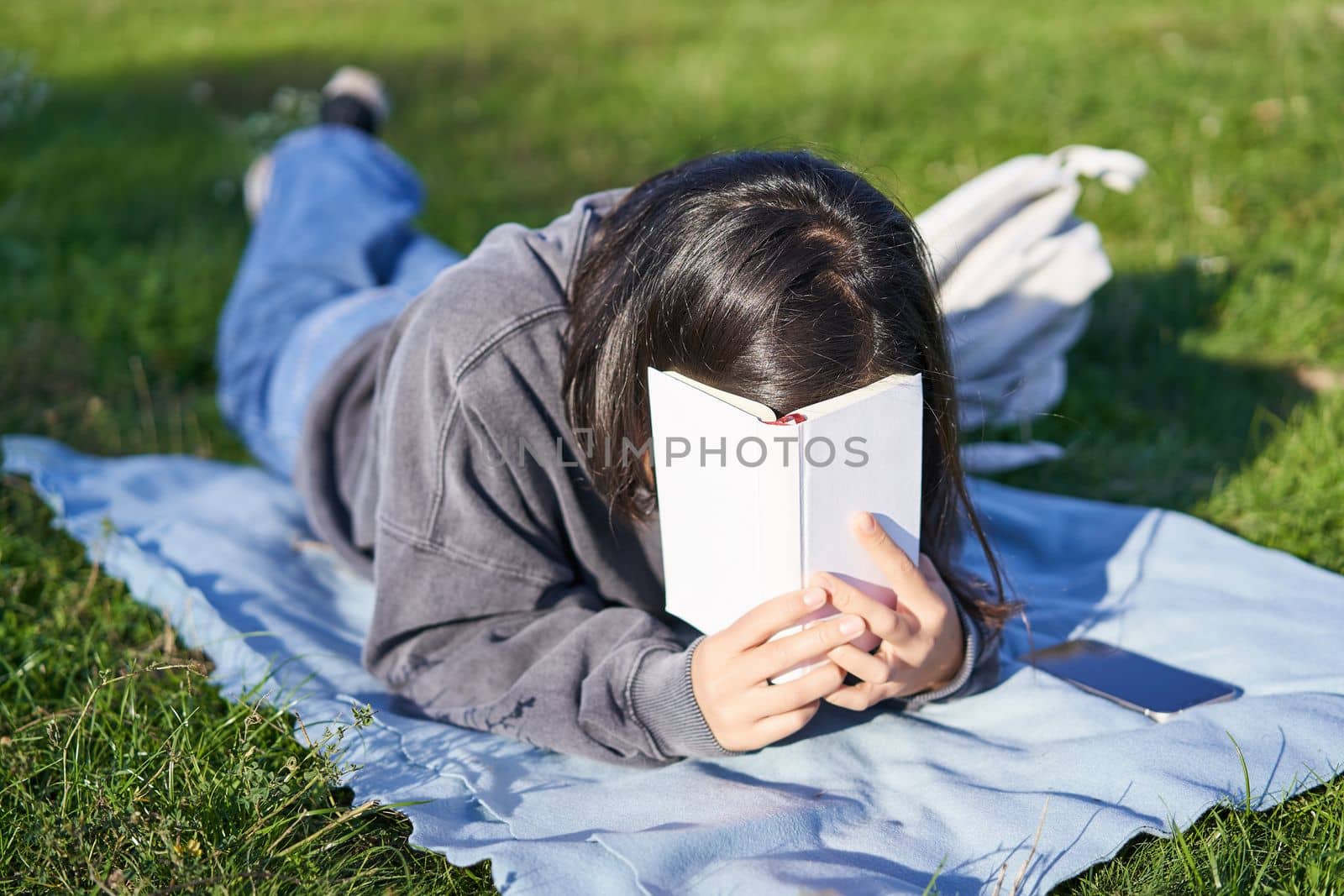 Portrait of happy girl laughing, covering face with her book, lying on grass in park. Copy space