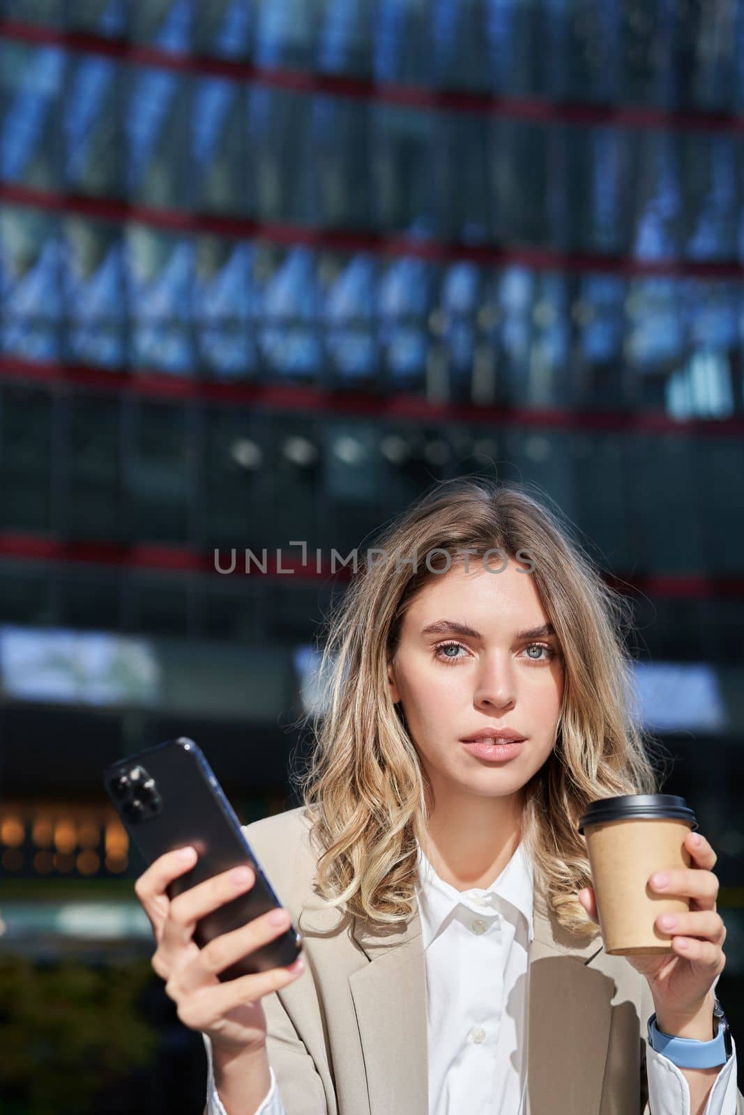 Stylish young corporate woman standing in street, drinks coffee and holds mobile phone.
