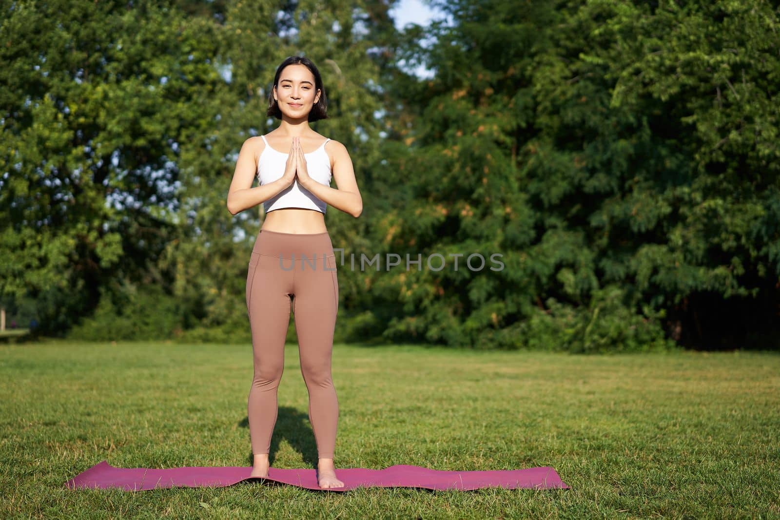 Young mindful woman, meditating on fresh air, doing yoga exercises in park, standing on rubber mat in sportswear by Benzoix