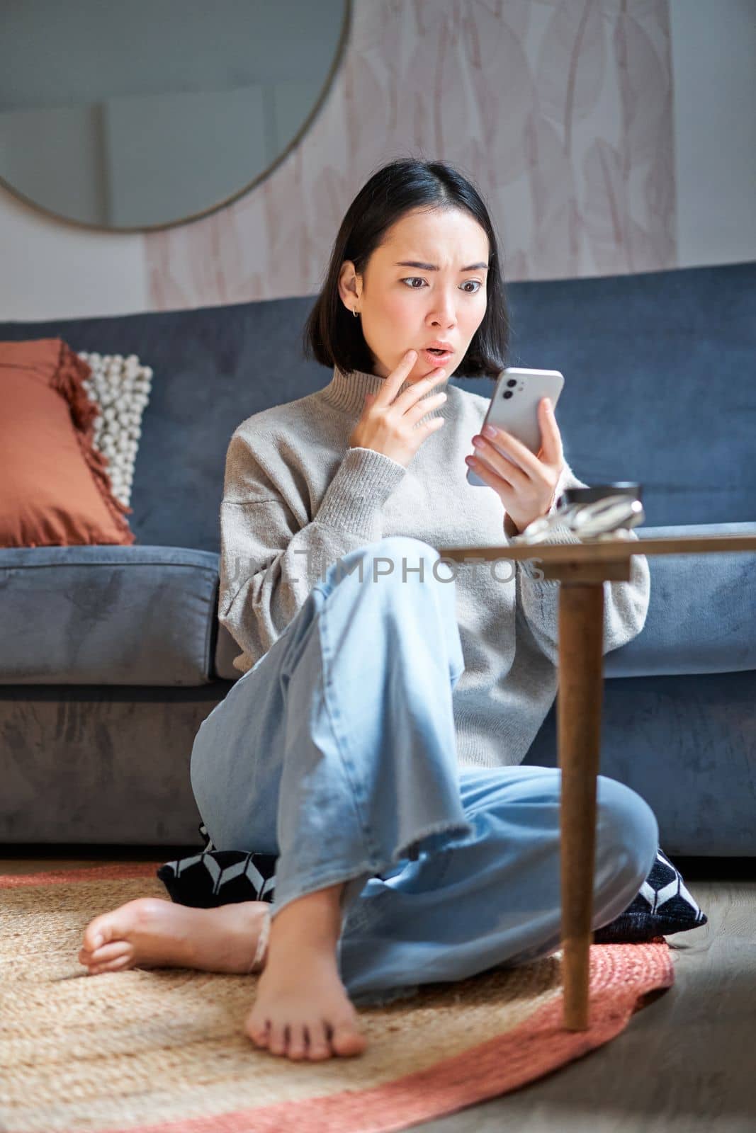 Vertical shot of young asian girl looks surprised at her mobile phone, reads amazing news, notification on smartphone app, sits at home on floor.