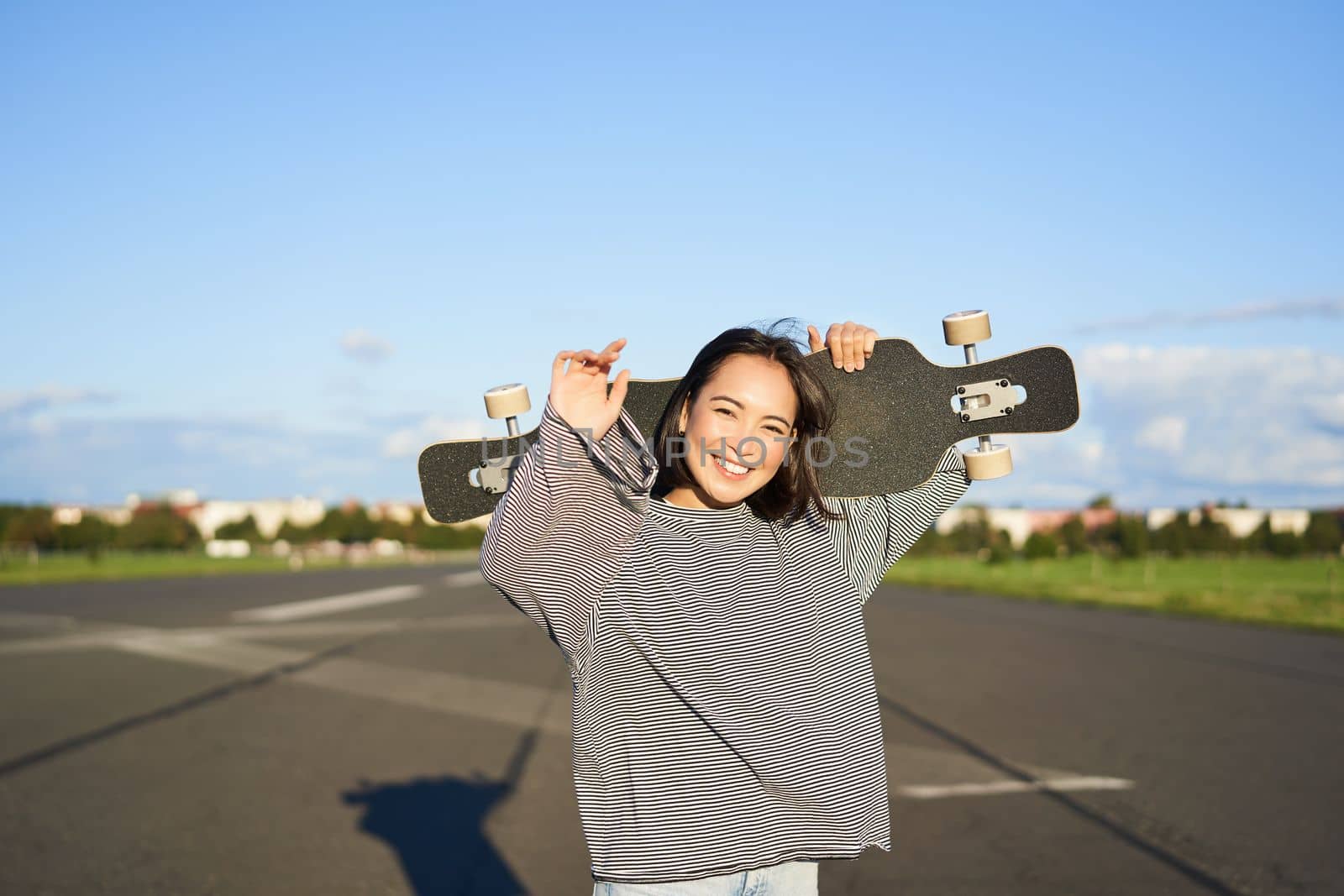 Leisure and people. Happy asian woman standing with longboard, cruising on an empty road in countryside. Skater girl holds her skateboard and smiles at camera by Benzoix