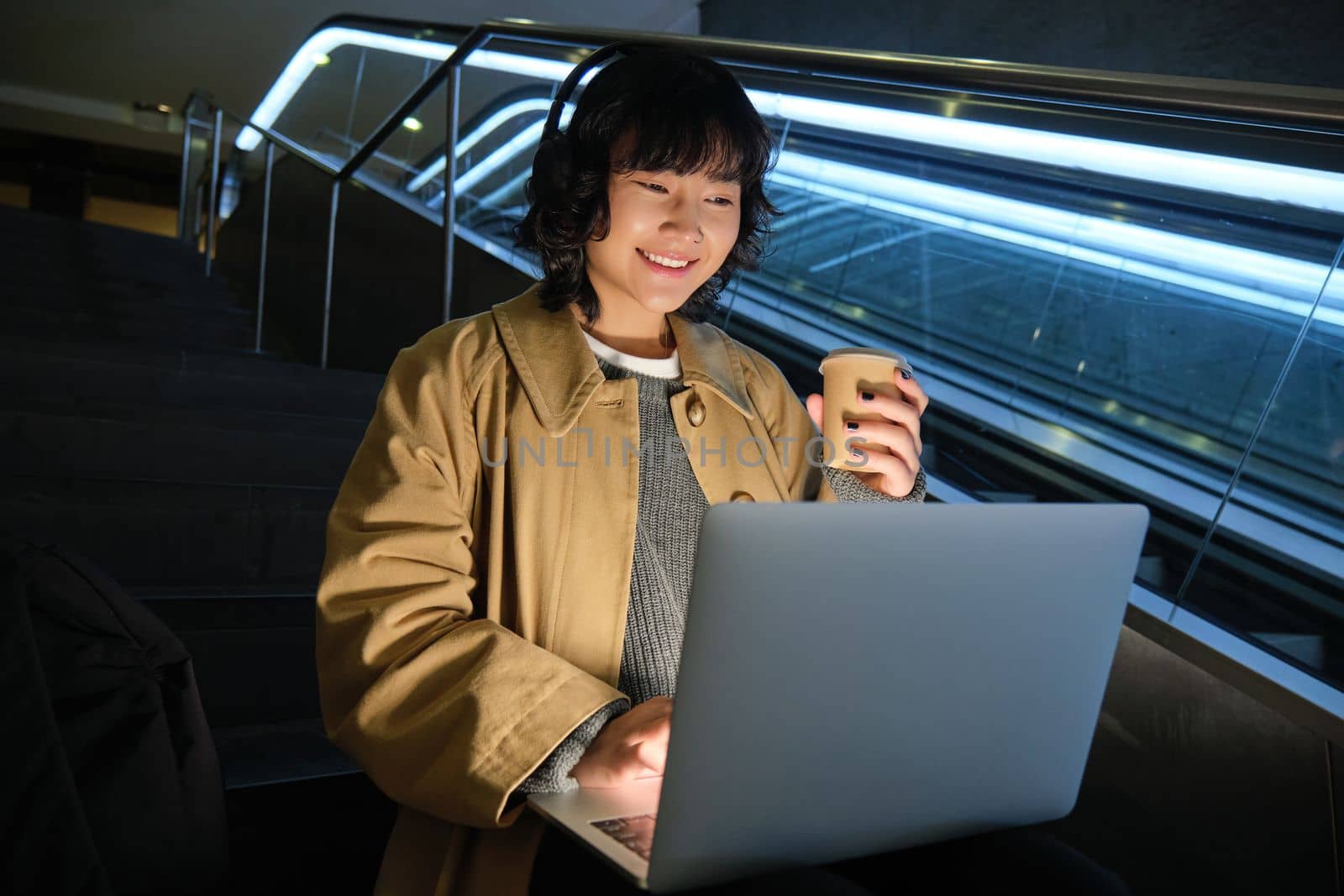 Young hipster girl in headphones, sits on staircase, drinks coffee and works on her laptop, studies in public place.