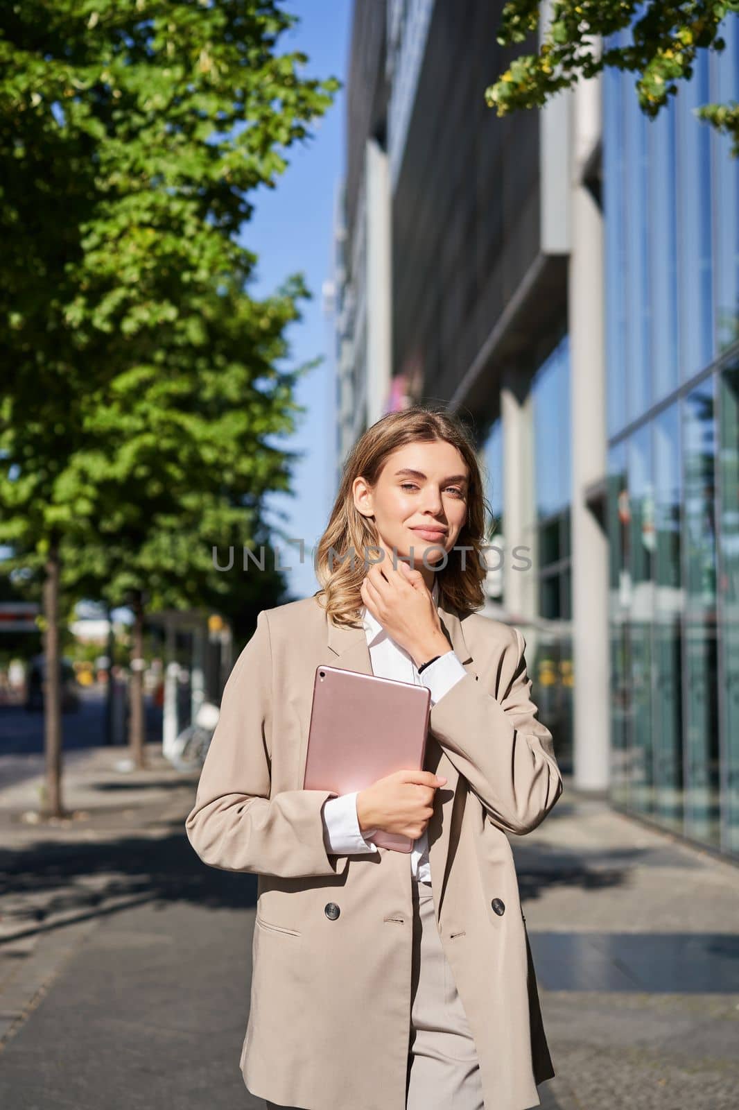 Vertical shot of successful business woman, team leader in suit, holding digital tablet, walking on street. Corporate lady goes to work by Benzoix