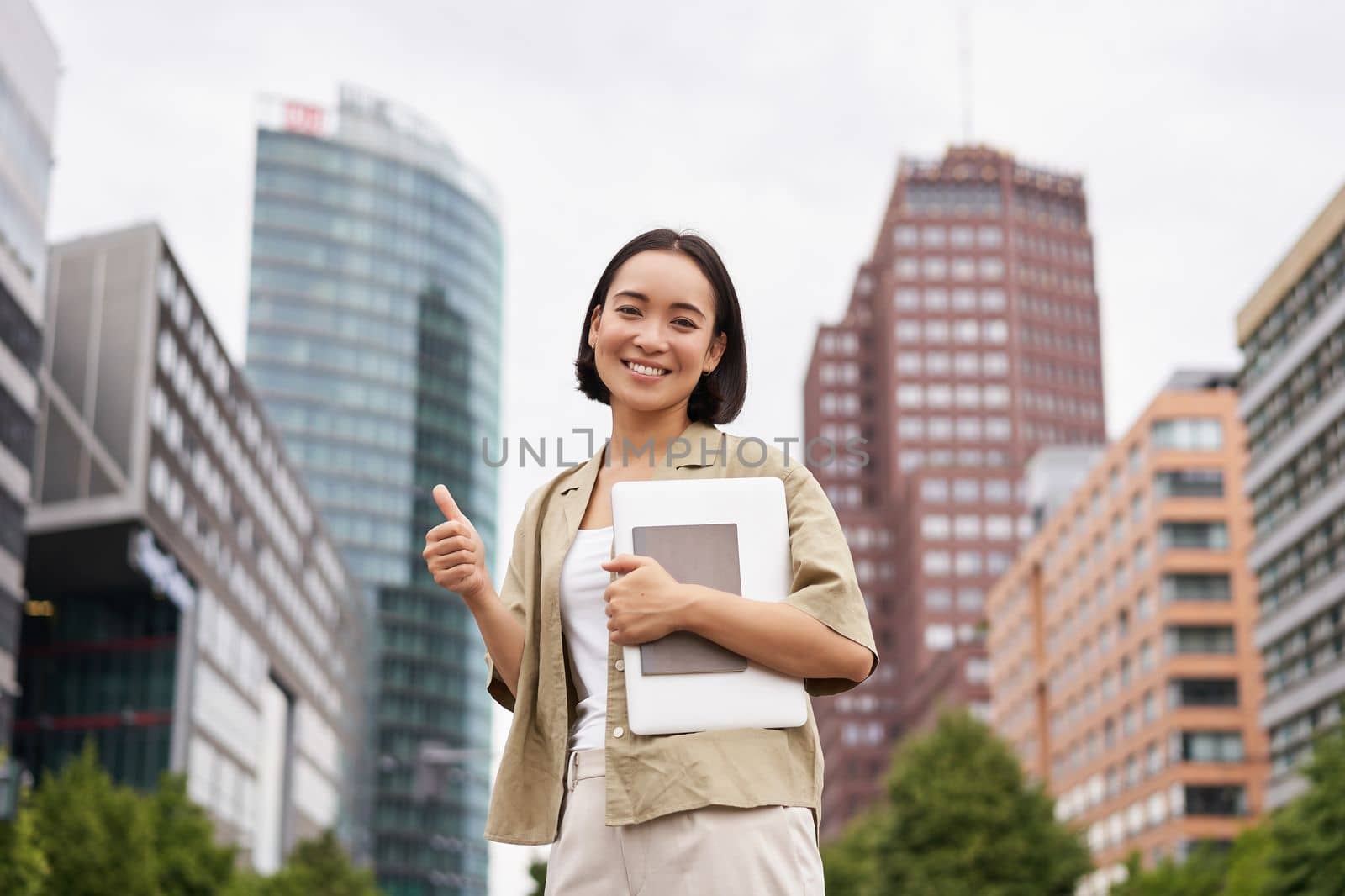 Cheerful asian girl in city centre, showing yes, hooray gesture, standing with laptop and celebrating.