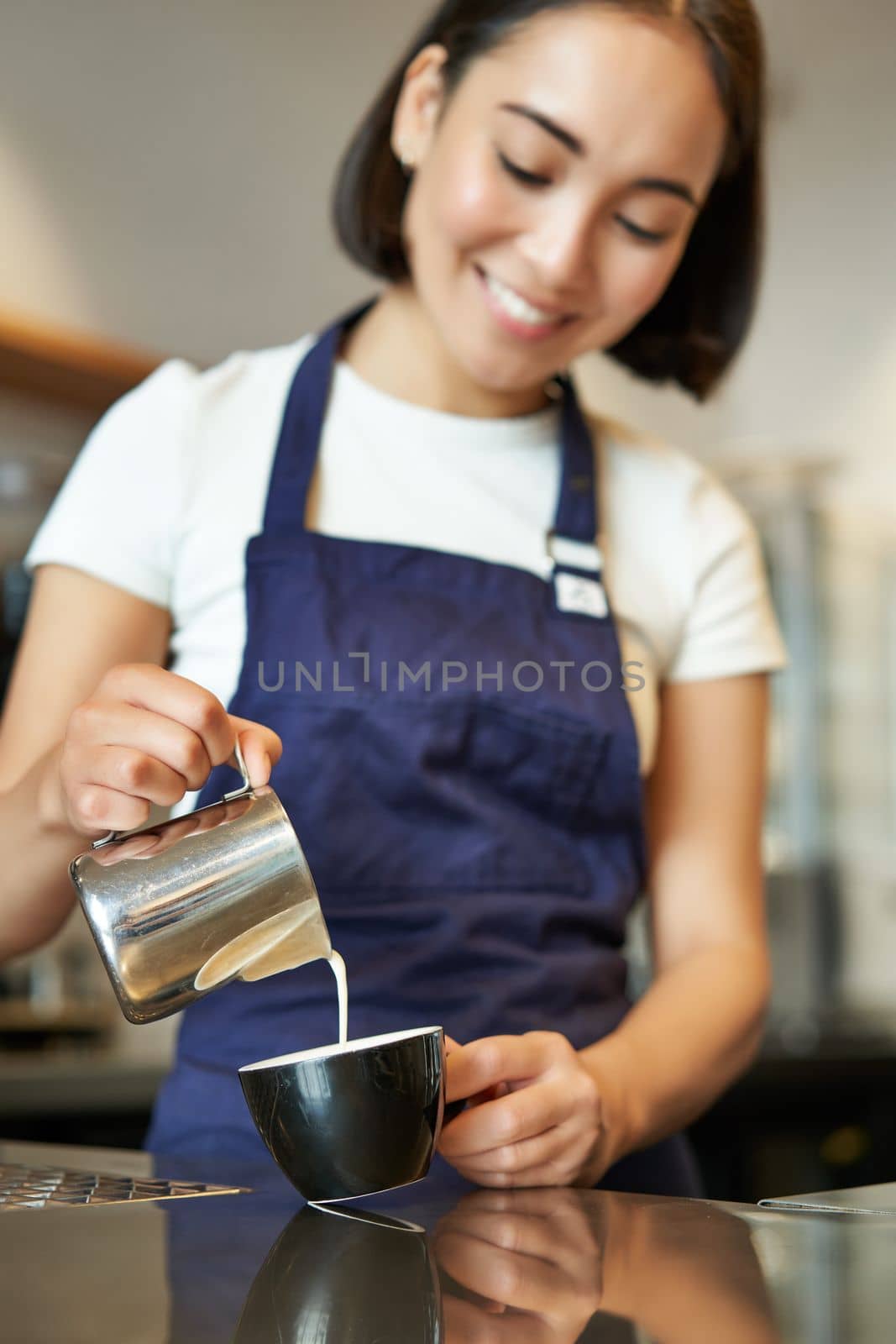 Vertical shot of brunette asian girl barista, wearing apron, making coffee, prepare order in cafe, pouring milk in cup for cappuccino latte art.