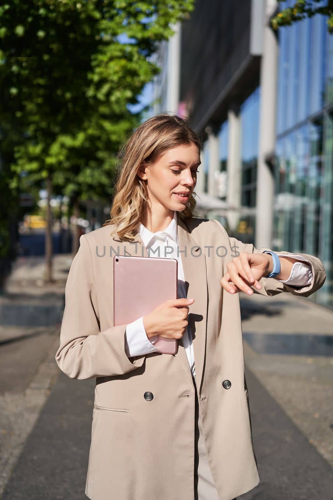 Vertical portrait of beautiful businesswoman in suit, holds her digital tablet, looks at watch on wrist, reads message or checks time.