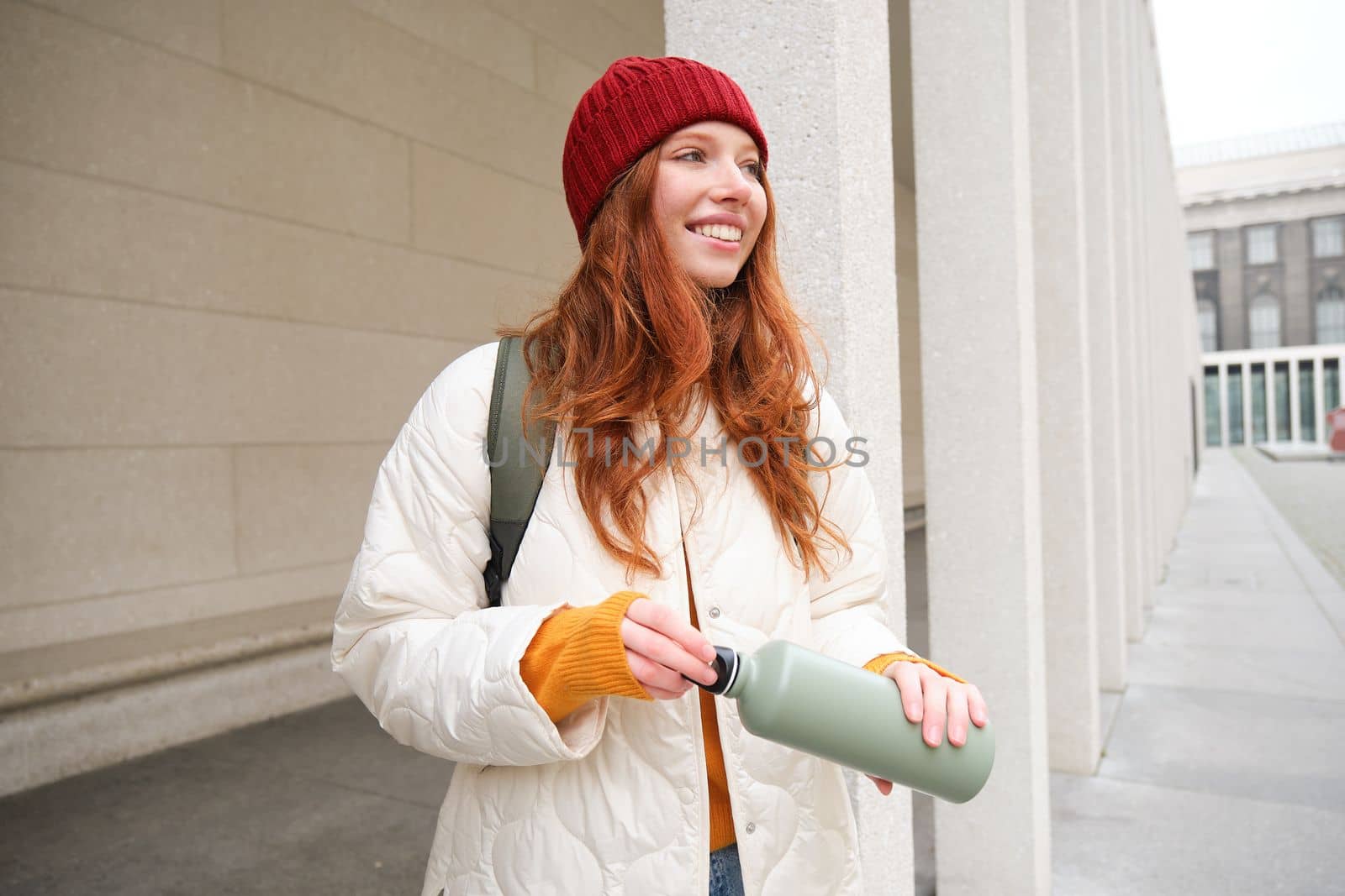 Young happy redhead woman in red hat, drinking from thermos, warming up with hot drink in her flask while walking around city, tourist relaxes with warm refreshemtn by Benzoix
