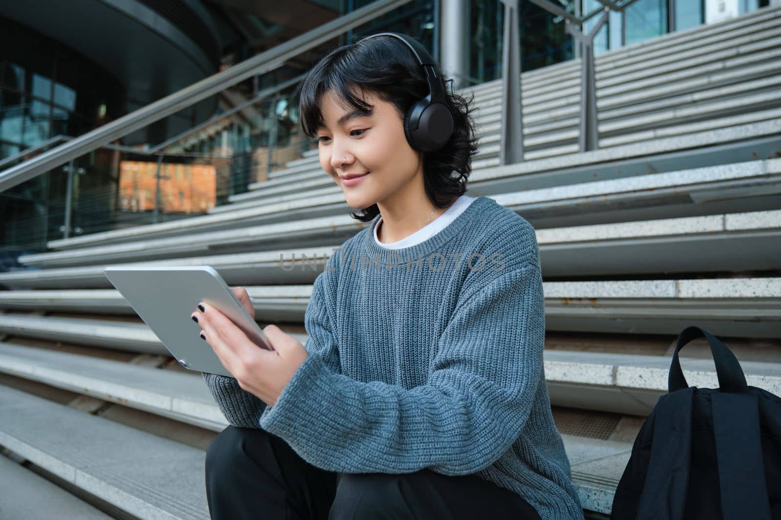 Smiling chinese student sits on stairs with tablet, draws digital art, graphic design project for freelance job, listens music in headphones and smiles by Benzoix