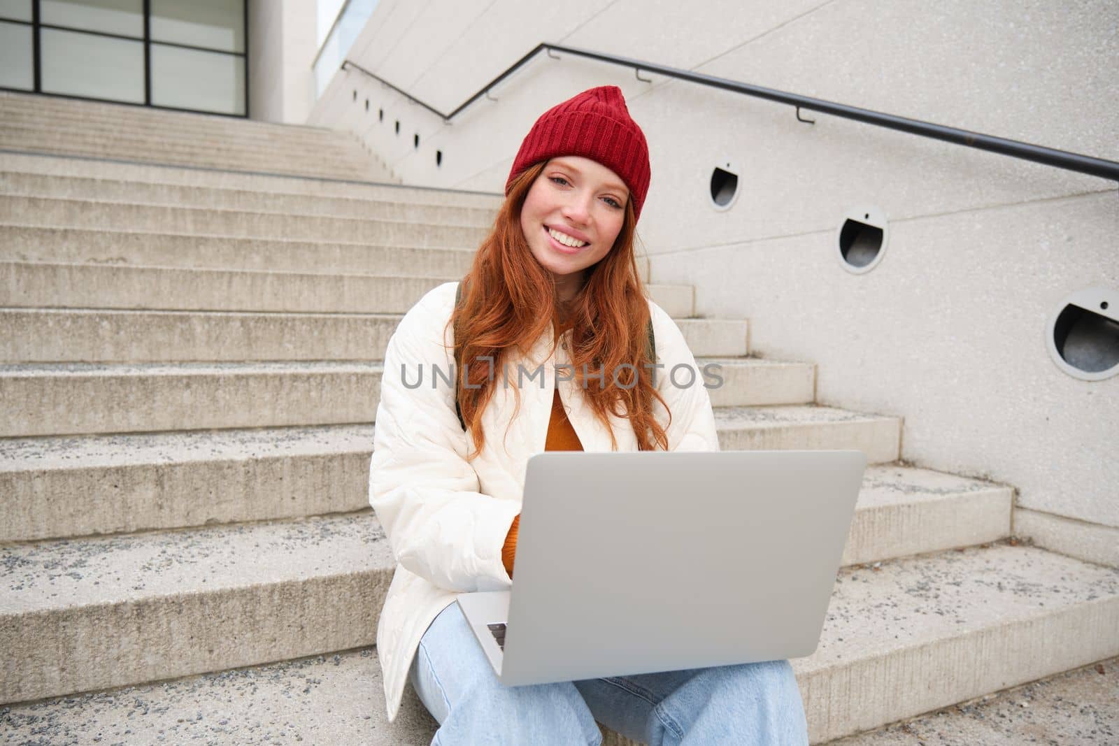 Smiling redhead girl, student sits on stairs outdoors and uses laptop, connects to public wifi in city and works on project, uses internet on computer by Benzoix