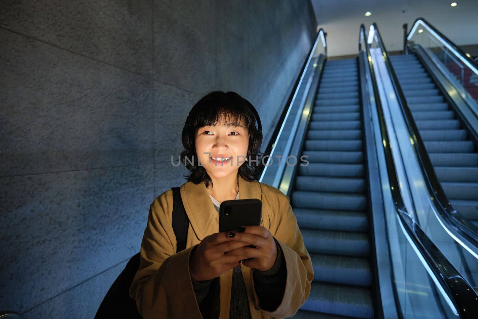 Cute young woman going down the escalator to the tube, using subway metro to commute to work or university, standing with smartphone.