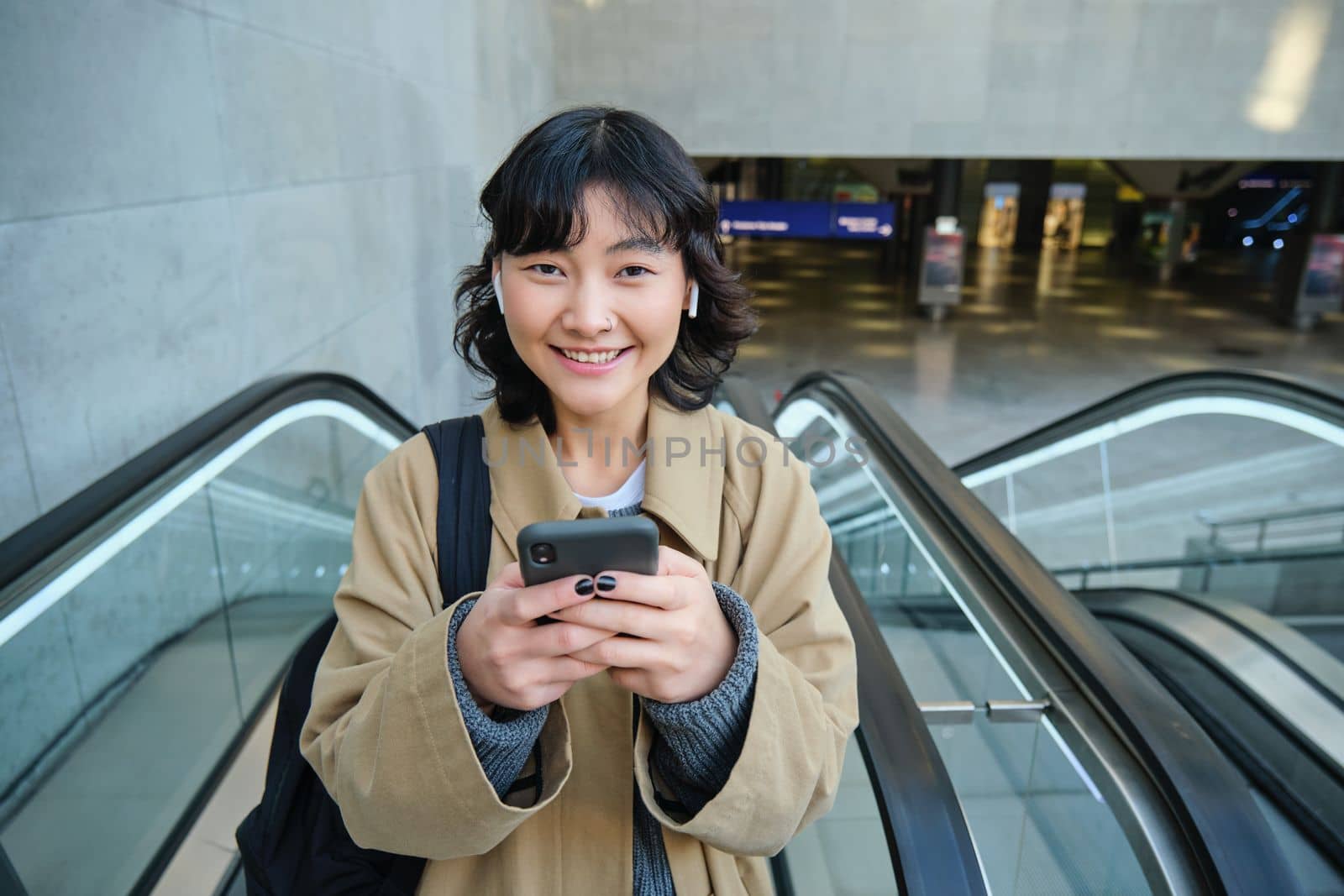 Portrait of cute smiling korean girl, goes up escalator, listens music in wireless earphones and uses mobile phone, holds smartphone.