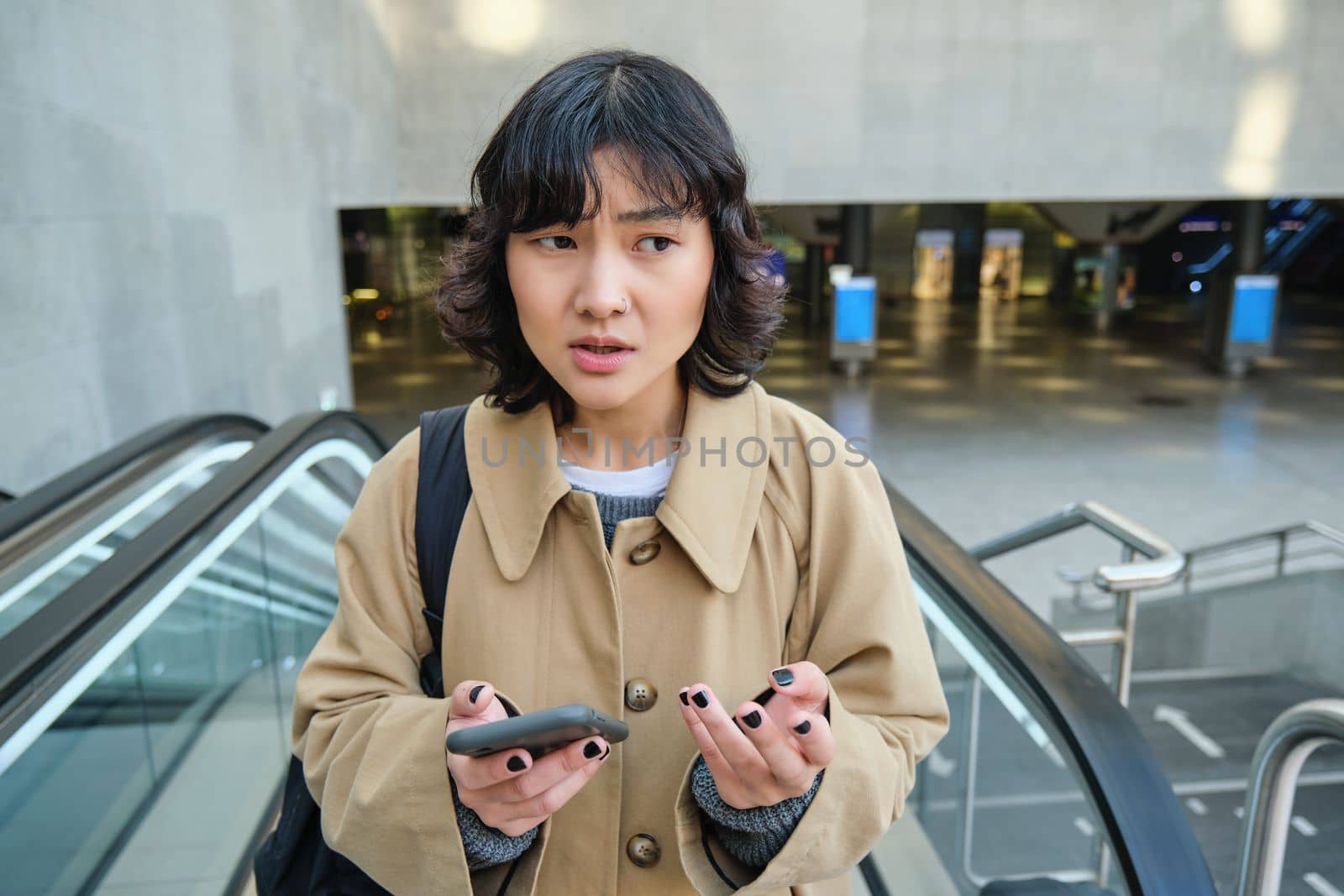 People in city. Portrait of korean girl standing on escalator, looks confused after reading text message on mobile phone.