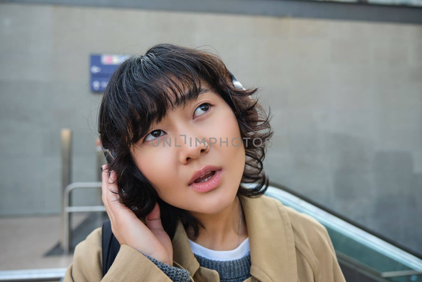 Portrait of stylish asian girl in headphones, listens podcast or music with confused face, looking up puzzled, seeing smth strange, standing on street.