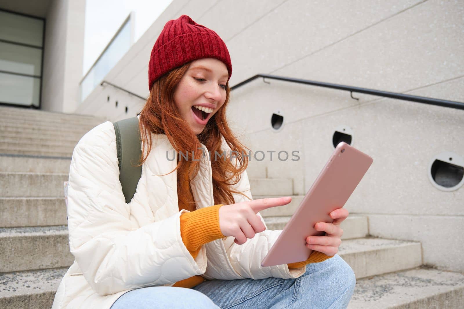 Young stylish girl, redhead female students sits on stairs outdoors with digital tablet, reads, uses social media app on gadget, plays games while waits on street.