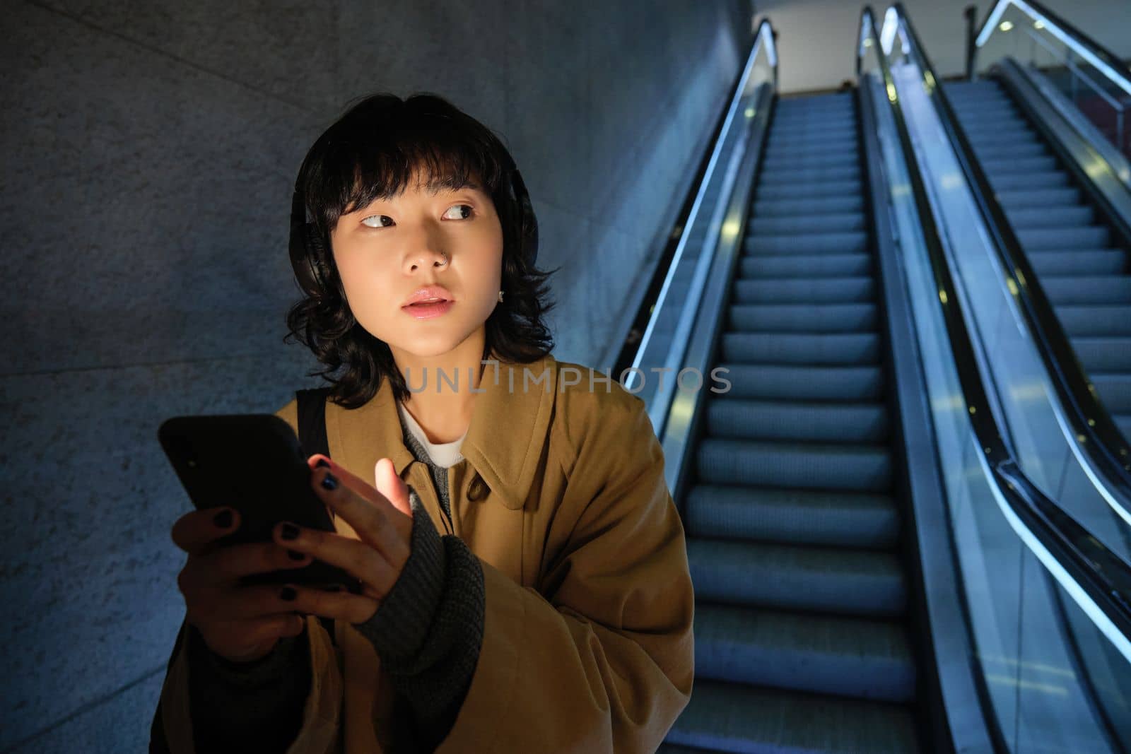 Urban lifestyle and people. Portrait of Korean girl in headphones, holds smartphone, looks concerned and tensed while going down escalator in dark by Benzoix